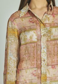 Camisa de lino con estampado que presenta suaves colores rosa, amarillo y crema, con un cuello tipo solapa, botones y diseños florales e intrincados.