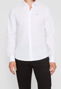 Camisa blanca de manga larga con botones y cuello puntiagudo, con el logo de CK en negro, combinada con pantalones negros de textura suave y tejida.