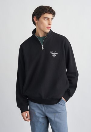 Jauns vīrietis valkā melnu ceturtdaļģerbijas džemperi ar "Carhartt WIP" logotipu, gaiši zilas bikses, stāv pret vienkāršu gaiši pelēku fonu.