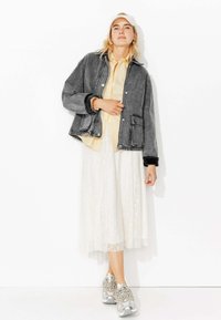 Veste en jean gris foncé à coupe décontractée, associée à une chemise rayée jaune clair et à une jupe en dentelle blanche. Des baskets argentées complètent le look.