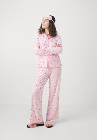Rosa sovklädset med röda hjärtan, långärmad, knäppbar skjorta och lösa byxor. Inkluderar en matchande rosa ögonmask.