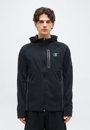 Νεαρός άνδρας με κοντά σγουρά μαλλιά, φορώντας μαύρο μπουφάν με κουκούλα και φερμουάρ, με τσέπη στο στήθος και λογότυπο στην αριστερή πλευρά.