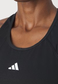 Gros plan sur le cou et le haut de la poitrine portant un haut de sport noir sans manches avec un petit logo triangulaire blanc centré sur la poitrine.