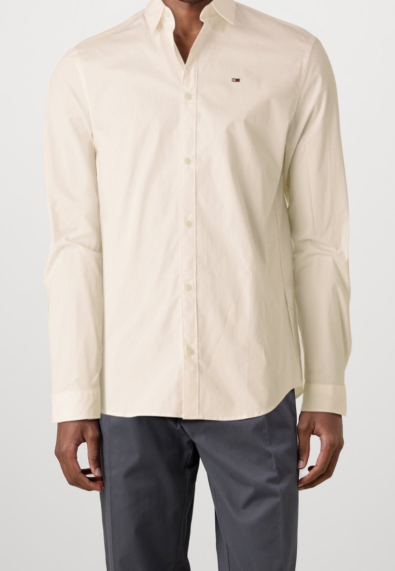 Homme portant une chemise crème à manches longues avec un petit logo sur la poitrine et un pantalon gris foncé, debout devant un fond uni.