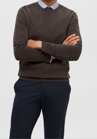 Brun stickad crewneck-tröja med ribbade mudd och nederkant, bärs över en ljusblå skjorta med krage, ihop med mörka skräddarsydda byxor.