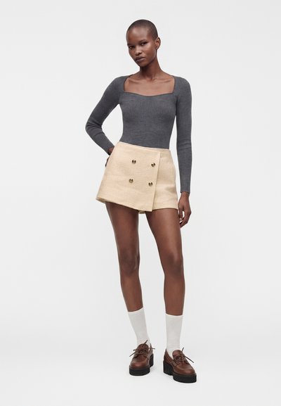 Femme portant un haut gris à manches longues, une jupe portefeuille beige boutonnée, des chaussettes blanches et des chaussures compensées marron, debout devant un fond blanc uni.