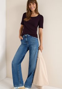 Burgunderfarbendes, eng anliegendes T-Shirt, kombiniert mit weit geschnittenen blauen Jeans. Das Model hält eine hellbeige Cordjacke. Klare Linien und lässige Passform.