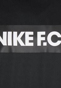 Schwarzes T-Shirt mit einer glatten Textur, das ein markantes weißes "NIKE F.C."-Logo in einem rechteckigen Streifen über der Brust zeigt.