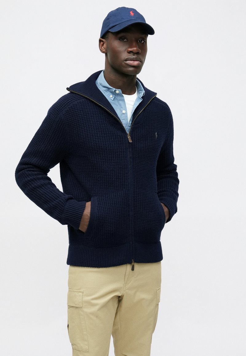 Suéter azul marino de punto con cremallera, patrón texturizado, puños acanalados y bolsillos. Combinado con una camisa azul clara y pantalones cargo beige.