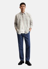 Karierter Hemd in Beige und Blau, kombiniert mit dunkelblauen Jeans. Das Model steht mit den Händen in den Taschen und trägt schwarze Schuhe.
