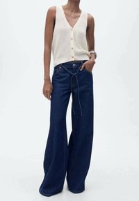 Osoba v krémovém bezrukávovém kardiganu na knoflíky a tmavě modrých širokých džínách s uvázaným opaskem, s jednou rukou v kapse na jednobarevném pozadí.