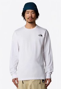 Wit T-shirt met lange mouwen van katoen, met een klein zwart logo op de borst, gecombineerd met een teal beanie en beige broek.