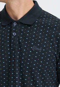 Polo noir avec fermeture à boutons, présentant un tissu texturé et un motif de points bleus et blancs, avec "Blend" brodé sur la poitrine.