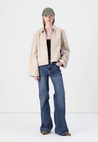 Beige læder cropped jakke med brede ærmer, åbnet, parret med blå udragende jeans og en grøn kasket. Ensfarvet baggrund.