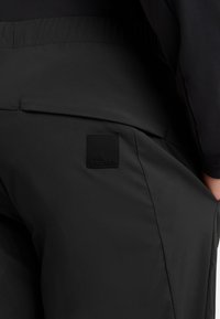 Pantalons noirs légers avec une taille élastique et une poche présentant une étiquette noire carrée embossée avec "Jack Wolfskin". Texture lisse.