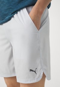 Persona vistiendo pantalones cortos ligeros grises de Puma con la mano en el bolsillo lateral, mostrando el logo de Puma cerca del dobladillo contra un fondo liso.