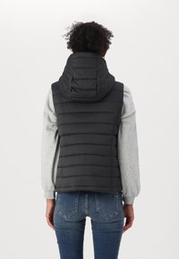 Doudoune noire avec capuche, présentant un design matelassé et une coupe ajustée, portée par-dessus un t-shirt à manches longues gris avec des poignets côtelés.