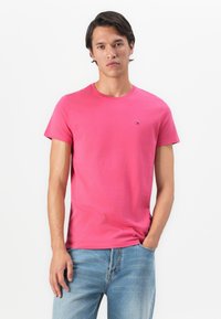 Homme portant un t-shirt rose à manches courtes et un jean bleu clair, se tenant avec une main dans la poche contre un fond blanc uni.