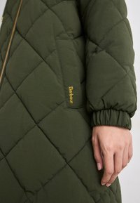 Gequiltede groene jas met een rits, voorzien van een opgestikte zak en elastische manchetten. "Barbour" is in het geel op de zak geborduurd.