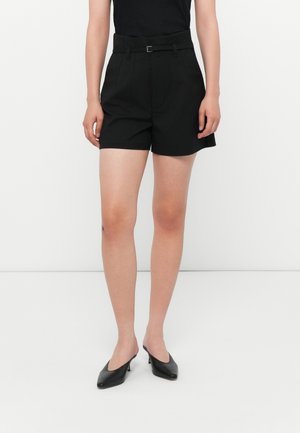 Shorts noirs ajustés avec une taille ceinturée, présentant une texture lisse et des plis marqués. Portés avec des escarpins noirs à talons ouverts.
