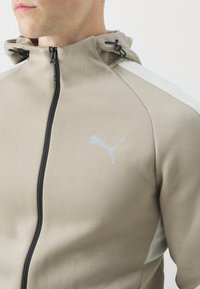 Giacca beige con zip e cappuccio, dotata di una zip nera, inserti testurizzati grigio chiaro sulle spalle e un logo Puma riflettente sul petto.