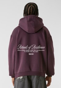 Lilla oversized hettegenser med snøring, ribbestrikkede mansjetter og hvit tekstdesign på baksiden. Stoffet virker mykt med en jevn finish.