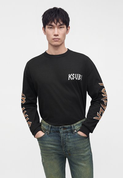 Jeune homme portant un t-shirt noir à manches longues avec le texte "KSUBI" sur la poitrine et un texte à motifs sur les manches, associé à un jean bleu délavé.