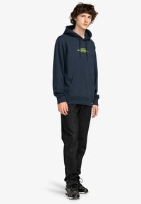 Navy blauwe hoodie met een voorzak en gele tekst. Gecombineerd met zwarte jeans en zwarte sneakers met een gestructureerde zool.