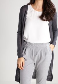 Cardigan lungo aperto di colore grigio scuro, realizzato in tessuto leggero. Abbinato a una maglietta bianca e pantaloni plissettati grigio chiaro.