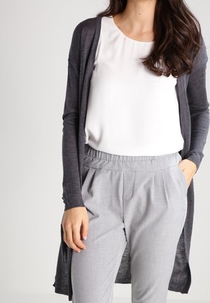 Cardigan lungo aperto di colore grigio scuro, realizzato in tessuto leggero. Abbinato a una maglietta bianca e pantaloni plissettati grigio chiaro.
