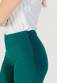 Pantaloni verdi a vita alta presentano una texture liscia, chiusura con zip laterale e una forma tailored. Abbinati a un top bianco aderente.