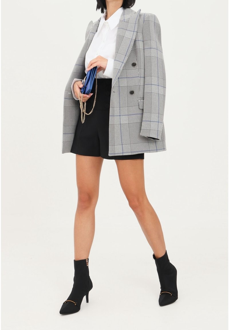 Blazer doppiopetto grigio a quadri sopra una camicia bianca, abbinato a pantaloni corti neri e stivaletti neri con tacco. Pochette blu con dettaglio a catena.