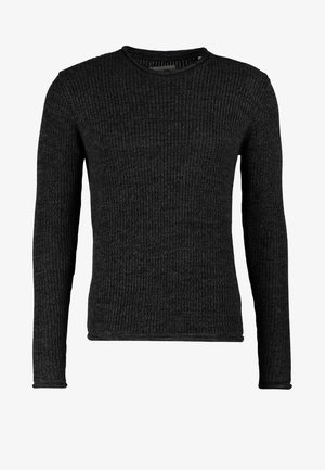 Uomo che cammina indossando un maglione nero, jeans scuri, sneakers blu navy e portando uno zaino nero su una spalla contro uno sfondo semplice.