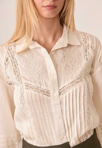 Blusa de lino, blanco roto con pliegues verticales y acentos de bordado floral. Presenta detalles de encaje y un cierre frontal con botones.