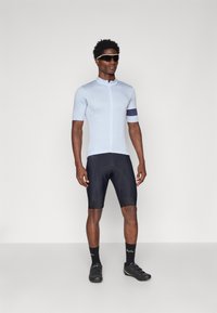 Tricou de ciclism bleu deschis cu mâneci scurte, închidere cu fermoar și bandă de culoare bleumarin la brațe, purtat cu pantaloni scurți cu pernițe negre și încălțăminte de ciclism negre.