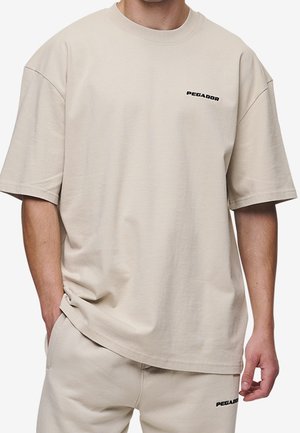 Beżowy bawełniany T-shirt o luźnym kroju, z krótkimi rękawami i małym czarnym logo „PEGADOR” na piersi. Prosty wzór bez żadnych ozdób.