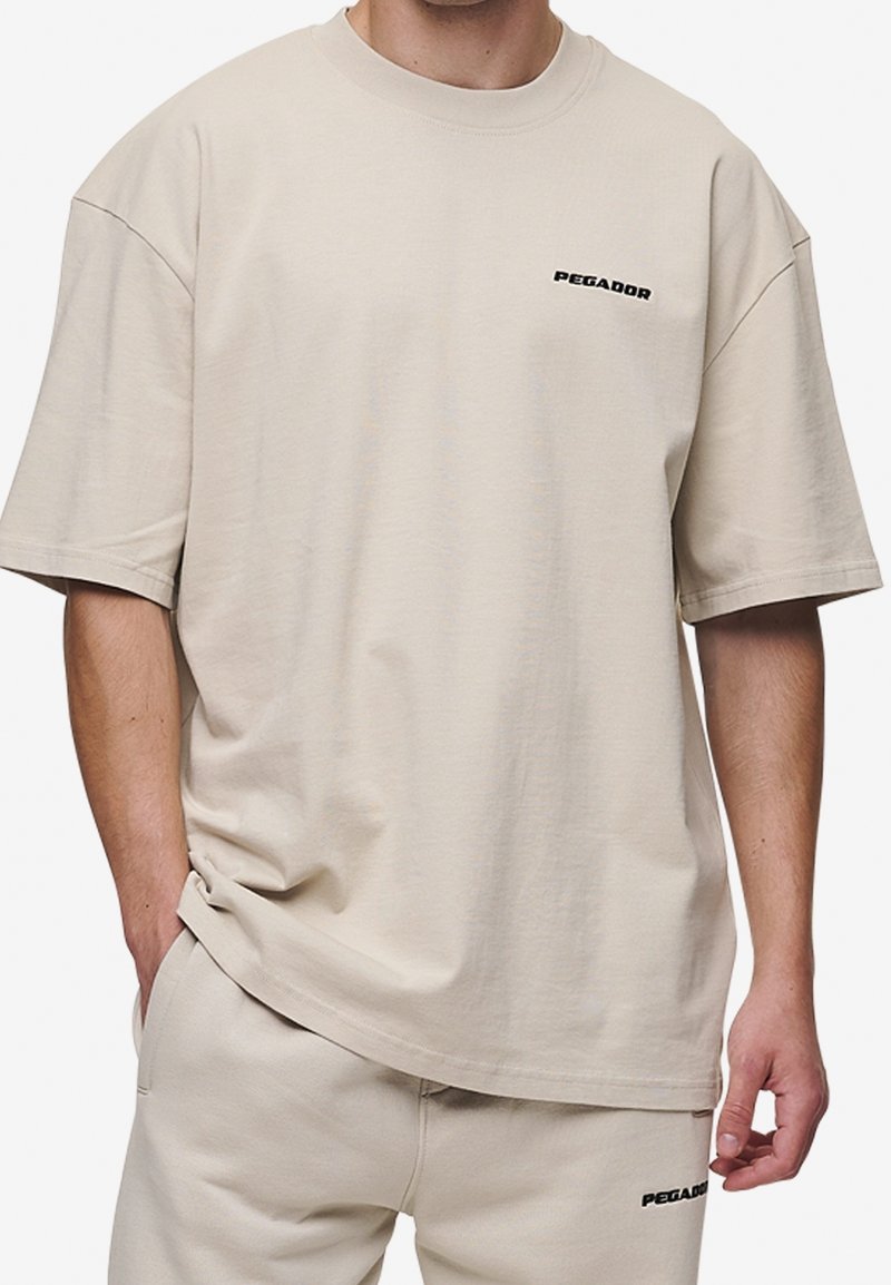 T-shirt beige en coton avec une coupe décontractée, manches courtes, et un petit logo noir "PEGADOR" sur la poitrine. Design uni sans motifs.