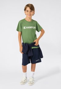 Žalias medvilninis marškinėliai su "CHAMPION" logotipu, tamsiai mėlynos šortai, tamsiai mėlynas džemperis, surištas aplink liemenį, balti kojinės ir smėlio spalvos sportbačiai su akcentais.