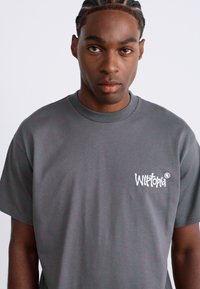 Jonge man met gevlochten haar, gekleed in een grijze T-shirt met een wit 'Wiltopia'-logo op de linkerborst, recht vooruit kijkend op een effen achtergrond.