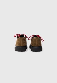 Brown suede shoes with chunky black soles, viewed from the back, featuring bright red laces tied in bows.