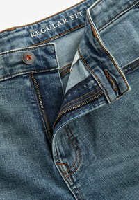 Jeans din denim cu o potrivire obișnuită, având o spălare de la albastru deschis la mediu, elemente de fixare de culoare cupru și detalii de cusătură vizibile.