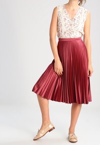 Roter, plissierter Midirock aus Satin, kombiniert mit einem ärmellosen Oberteil in hellen Farben mit Blumenmuster. Tragen Sie dazu beiges, flaches Schuhwerk.