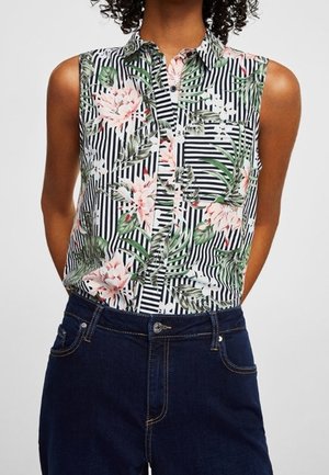 Femme portant un chemisier sans manches à rayures noires et blanches avec un motif floral vert et rose, associé à un jean bleu foncé.