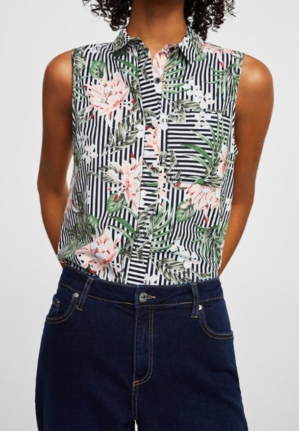 Femme portant un chemisier sans manches à rayures noires et blanches avec un motif floral vert et rose, associé à un jean bleu foncé.