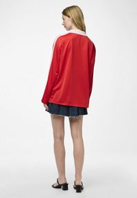 Rote Langarmjacke mit weißen Akzenten, präsentiert über einem Jeansrock. Glatter Stoff, lockere Passform, Kragen. Schlicher Hintergrund.