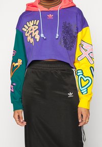 Felpa cropped viola con cappuccio rosa e maniche verdi e gialle. Caratterizzata da grafiche colorate. Abbinata a una gonna in satin nera, logo Adidas visibile.