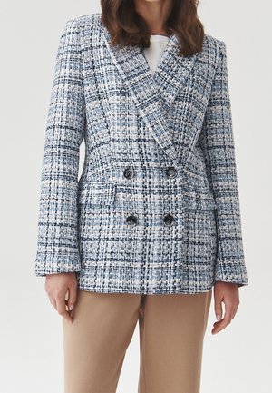 Femme portant un blazer croisé à carreaux bleu et blanc avec des boutons noirs et un pantalon beige sur un fond uni.