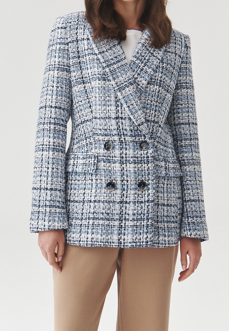 Femme portant un blazer croisé à carreaux bleu et blanc avec des boutons noirs et un pantalon beige sur un fond uni.