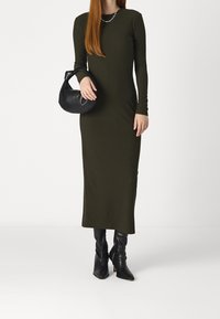 Olivově zelené žebrované šaty s dlouhým rukávem a přiléhavým střihem, v kombinaci s černou kabelkou a černými špičatými kotníkovými botami.