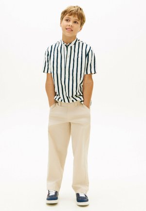 Ragazzo con le mani in tasca che indossa una camicia a maniche corte a righe verticali, pantaloni beige e sneakers blu navy, in piedi davanti a uno sfondo bianco.